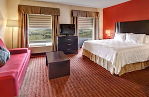 A hotel room with a bed, a TV on a dresser, a red sofa, and a view of the countryside.