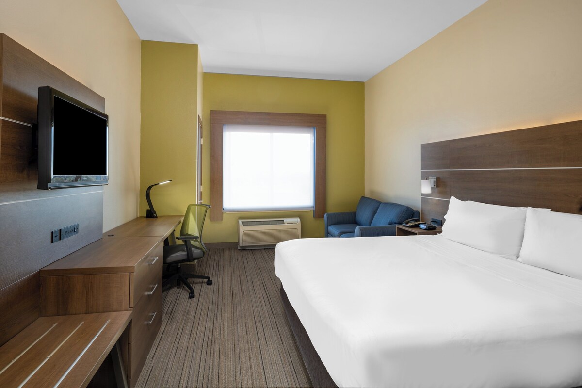 A hotel room with a bed, a desk with a TV, a chair, a window, and a nightstand.