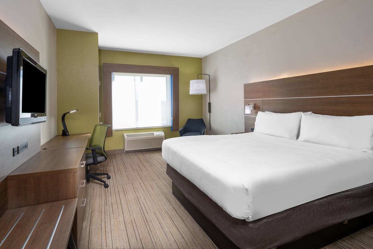 A hotel room with a large bed, a desk with a computer, a chair, a TV, and a window with natural light.