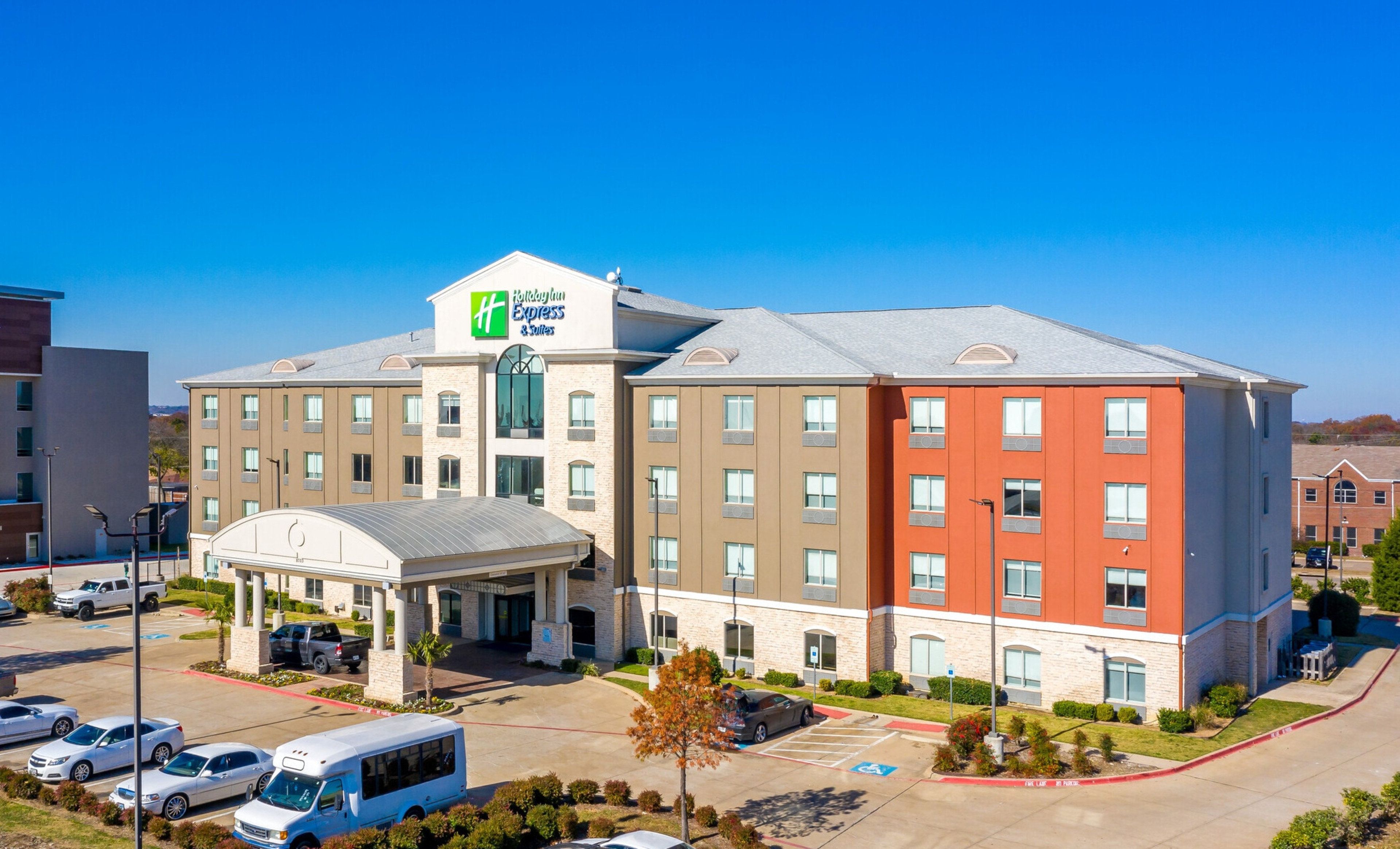 A multi-story hotel with a central entrance, parking lot, and a clear blue sky.