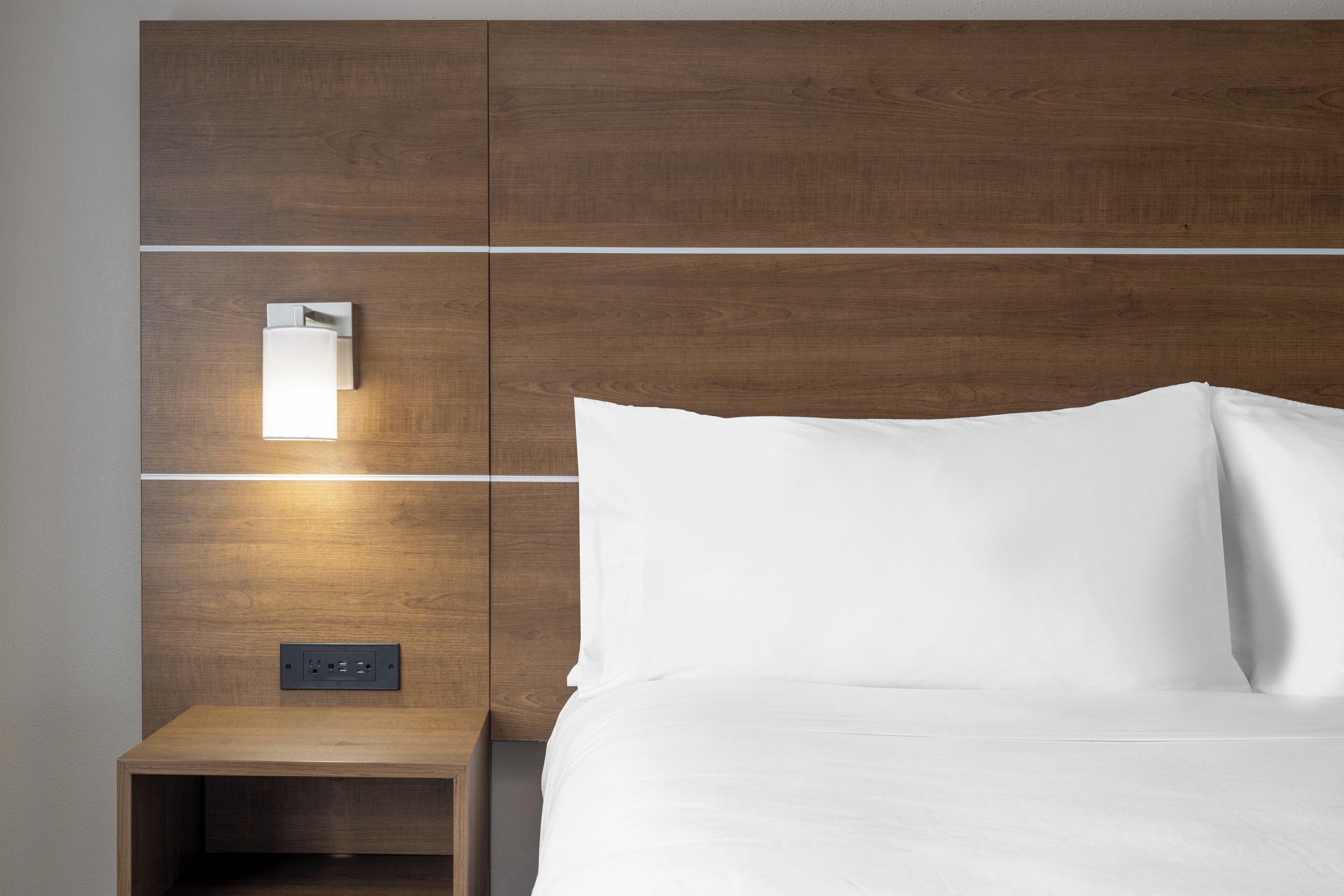 Een hotelkamer met een houten hoofdeinde, een nachtkastje met een lamp, en een bed met witte beddengoed.