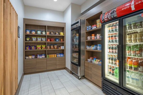 A snack and beverage vending area with a refrigerator and shelves stocked with snacks and drinks. - Holiday Inn Express & Suites Dallas East - Fair Park by IHG (Dallas)