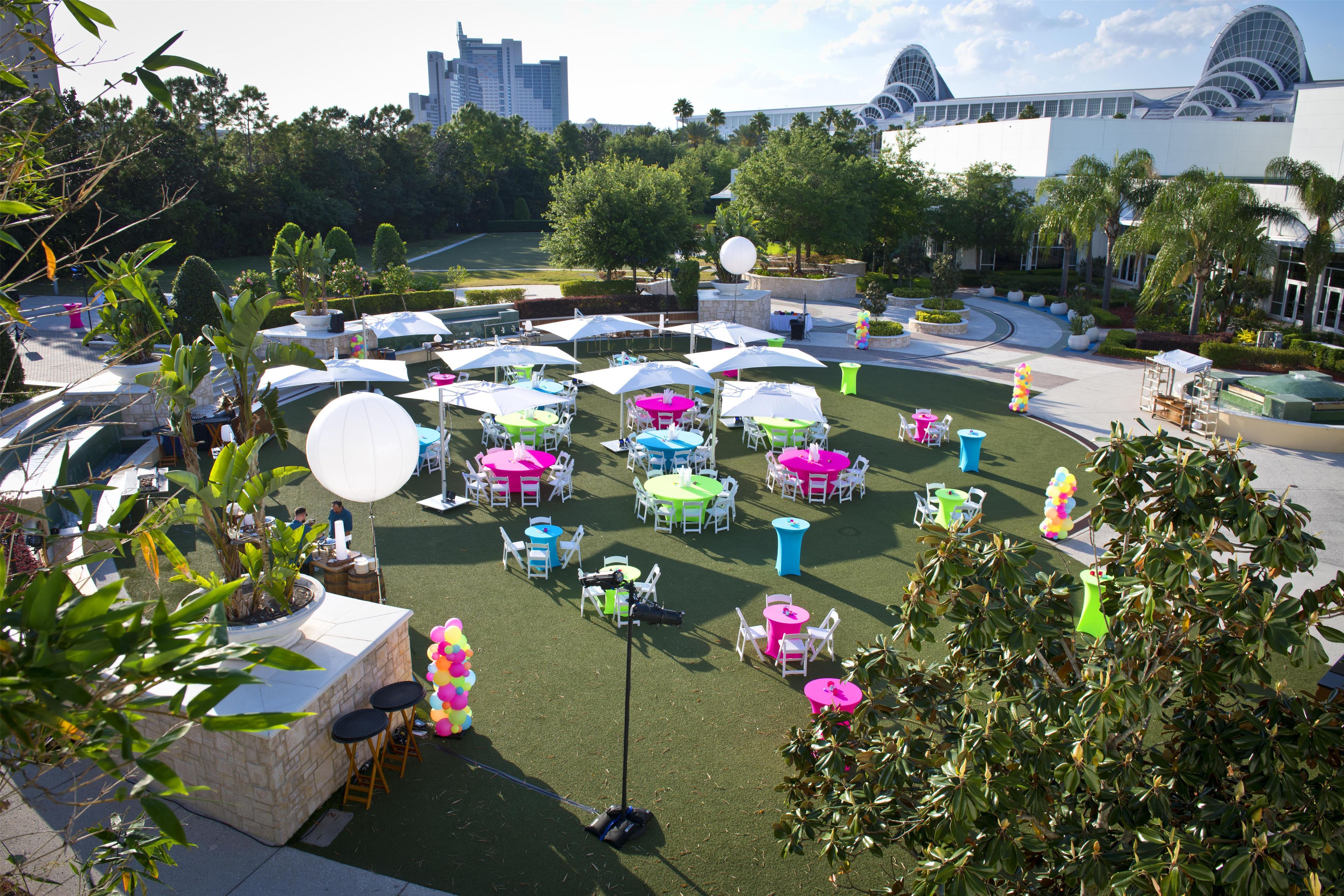 An outdoor event space with multiple tables and chairs, surrounded by greenery and a building in the background.