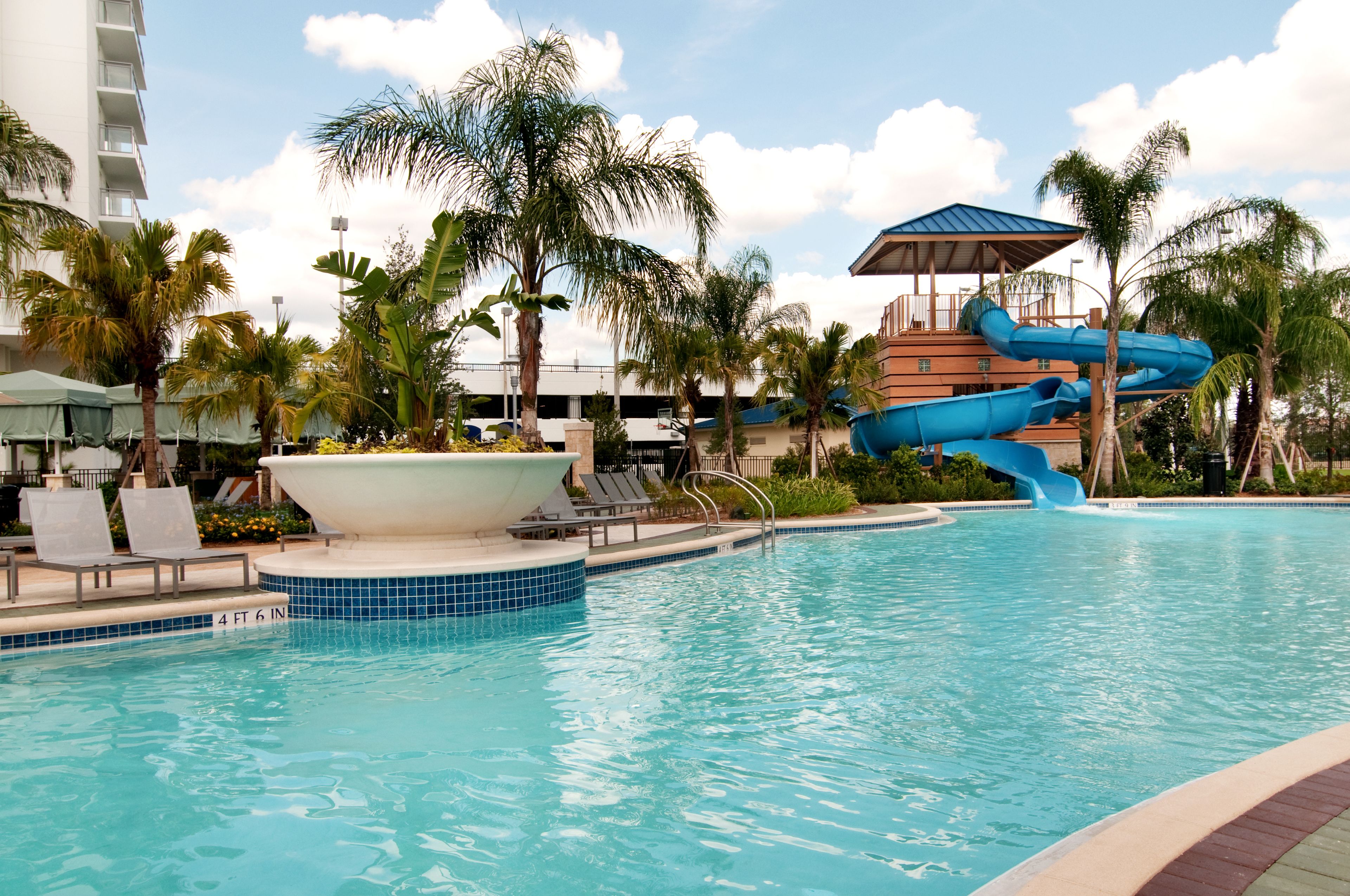A swimming pool with a water slide, lounge chairs, palm trees, and a building with a blue roof.