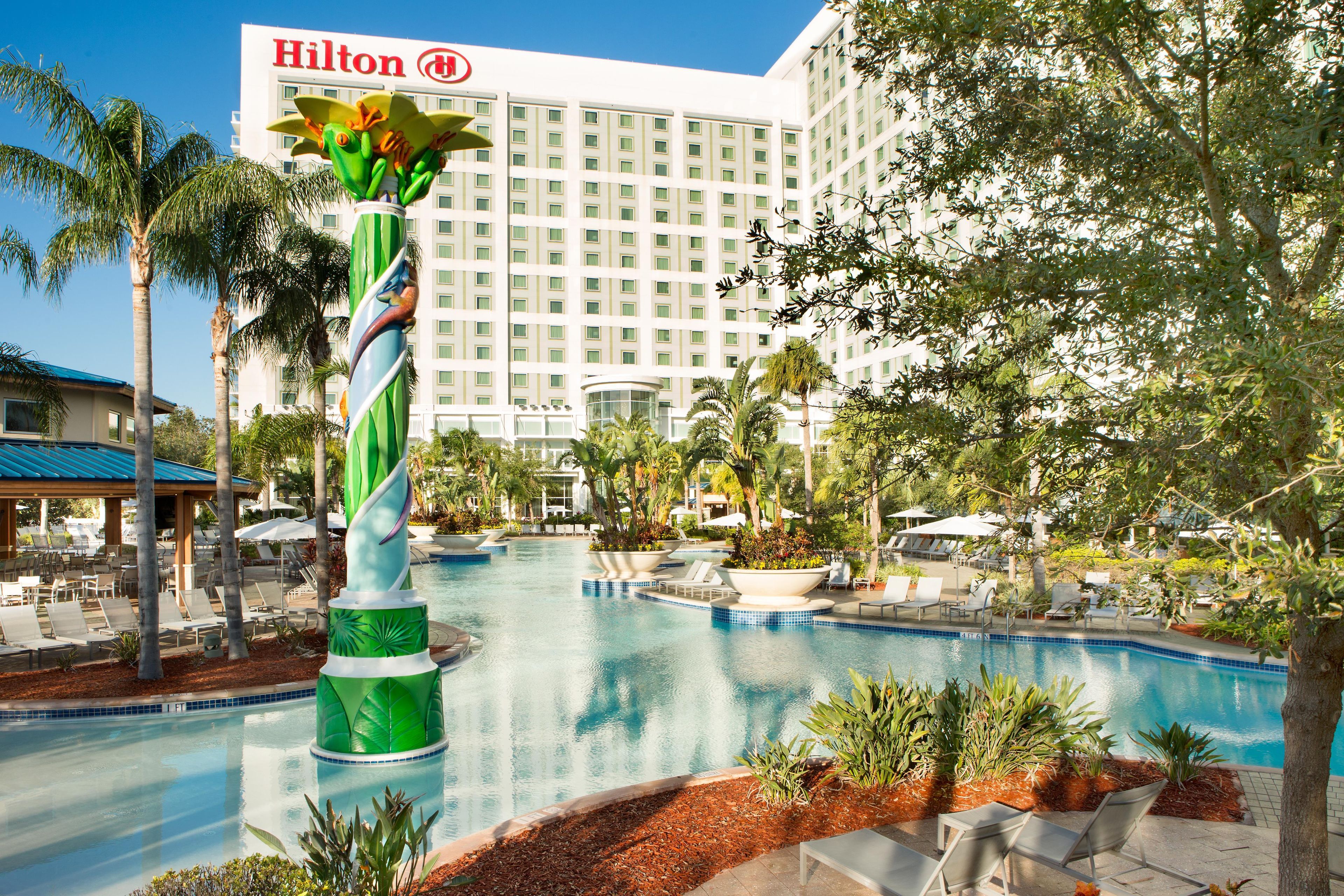 A swimming pool with lounge chairs, a large green decorative structure, and a Hilton hotel in the background.