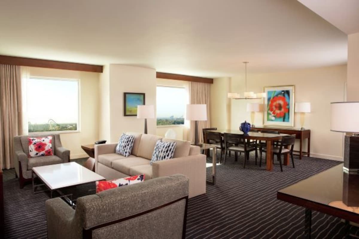 Habitación de hotel con sofá, sillón, mesa de comedor y sillas. Tiene ventanas con cortinas, un cuadro en la pared y una lámpara.