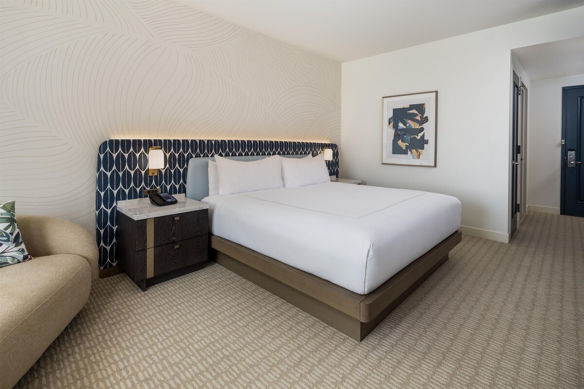 Habitación de hotel con cama, mesita de noche, lámpara, un cuadro en la pared y una puerta.