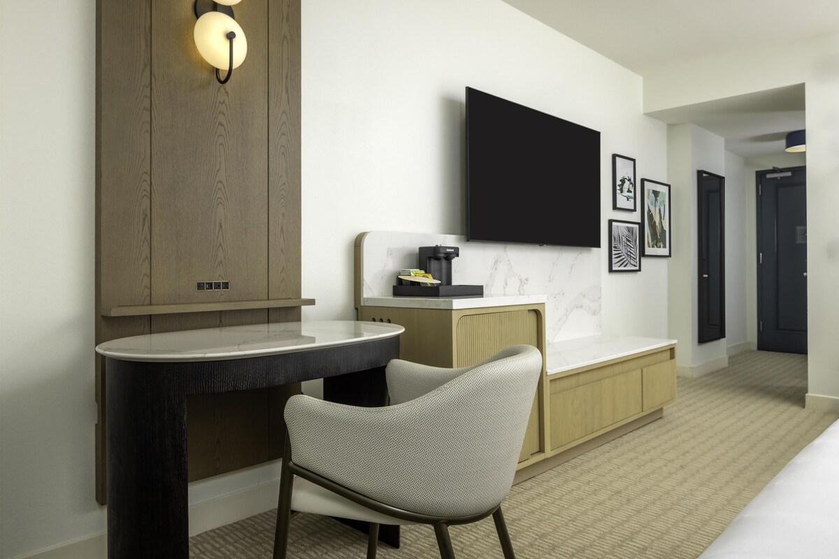 Una habitación de hotel moderna con una televisión de pantalla plana, una mesa redonda, una silla, un minibar y un armario de madera.