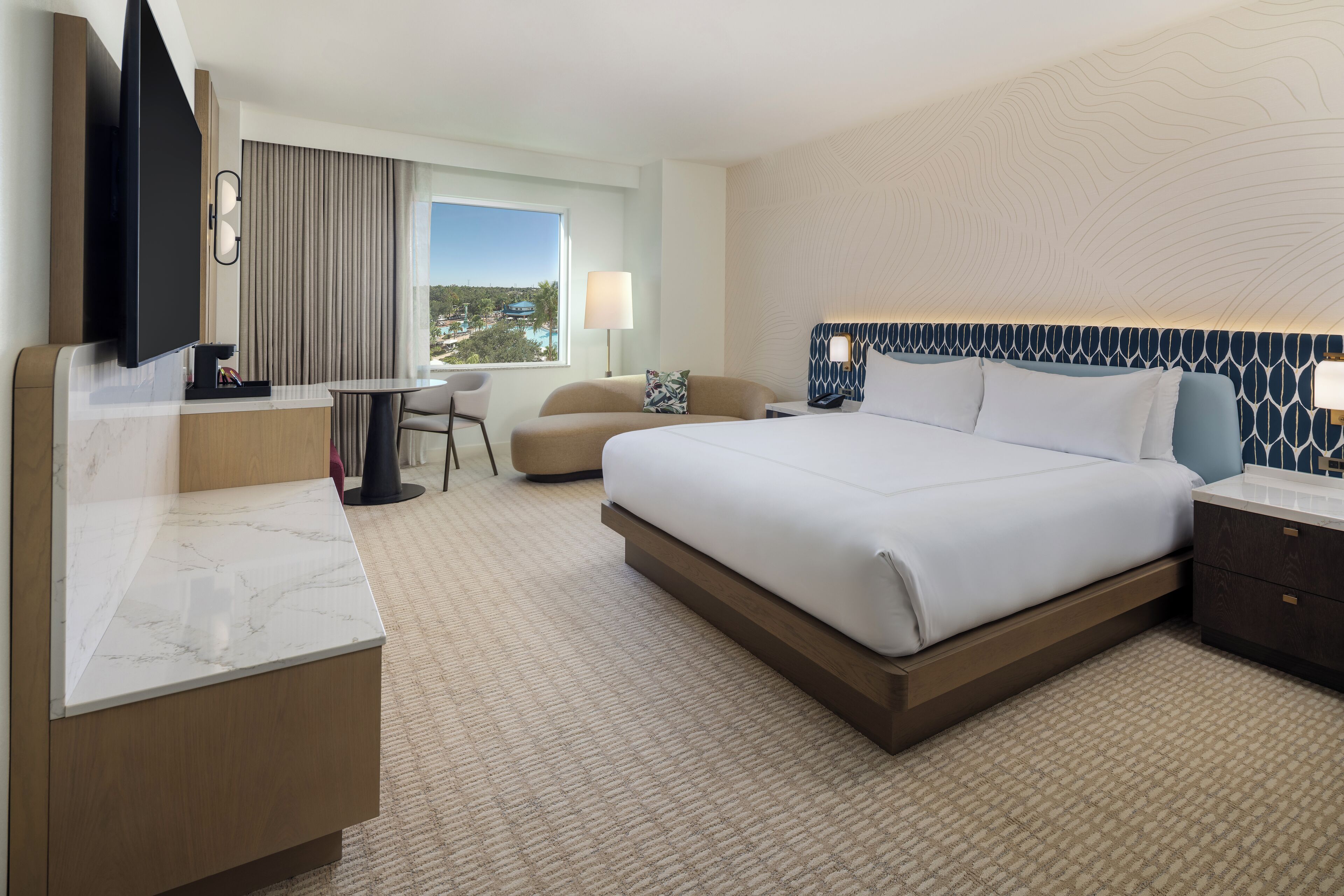 A hotel room with a large bed, a desk with a flat-screen TV, a chair, a small table, and a view of the city.