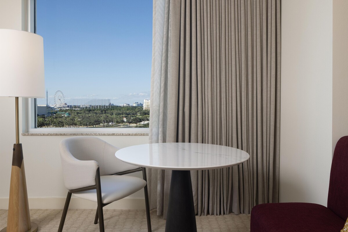 Habitación de hotel con una mesa redonda, una silla, una lámpara y vistas a un paisaje urbano a través de un ventanal amplio.