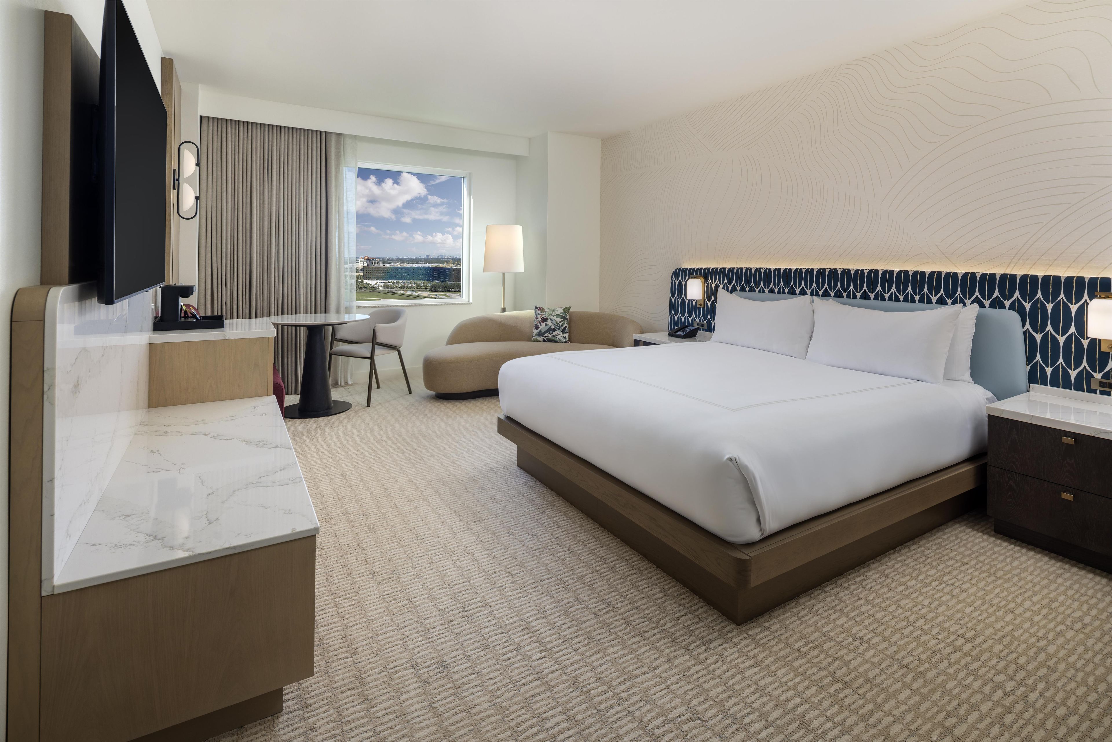 A hotel room with a large bed, a desk with a flat-screen TV, a chair, a lamp, and a view of the city.
