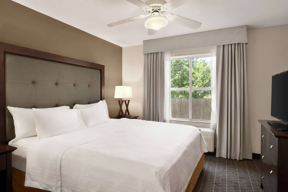 Une chambre d’hôtel avec un grand lit, un ventilateur de plafond, une table de chevet avec une lampe, un téléviseur posé sur une commode et une fenêtre avec des rideaux.