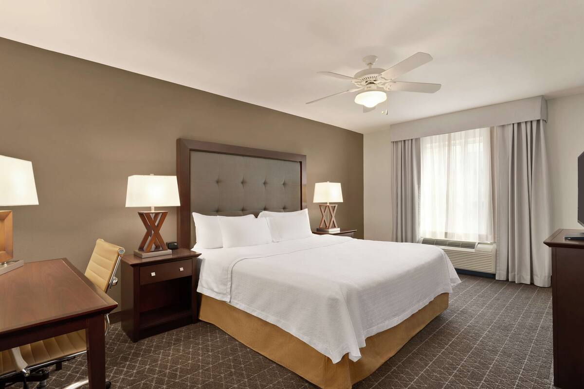 Une chambre d’hôtel avec un grand lit, deux tables de chevet avec des lampes, un bureau, une chaise, un ventilateur de plafond et une grande fenêtre avec des rideaux.