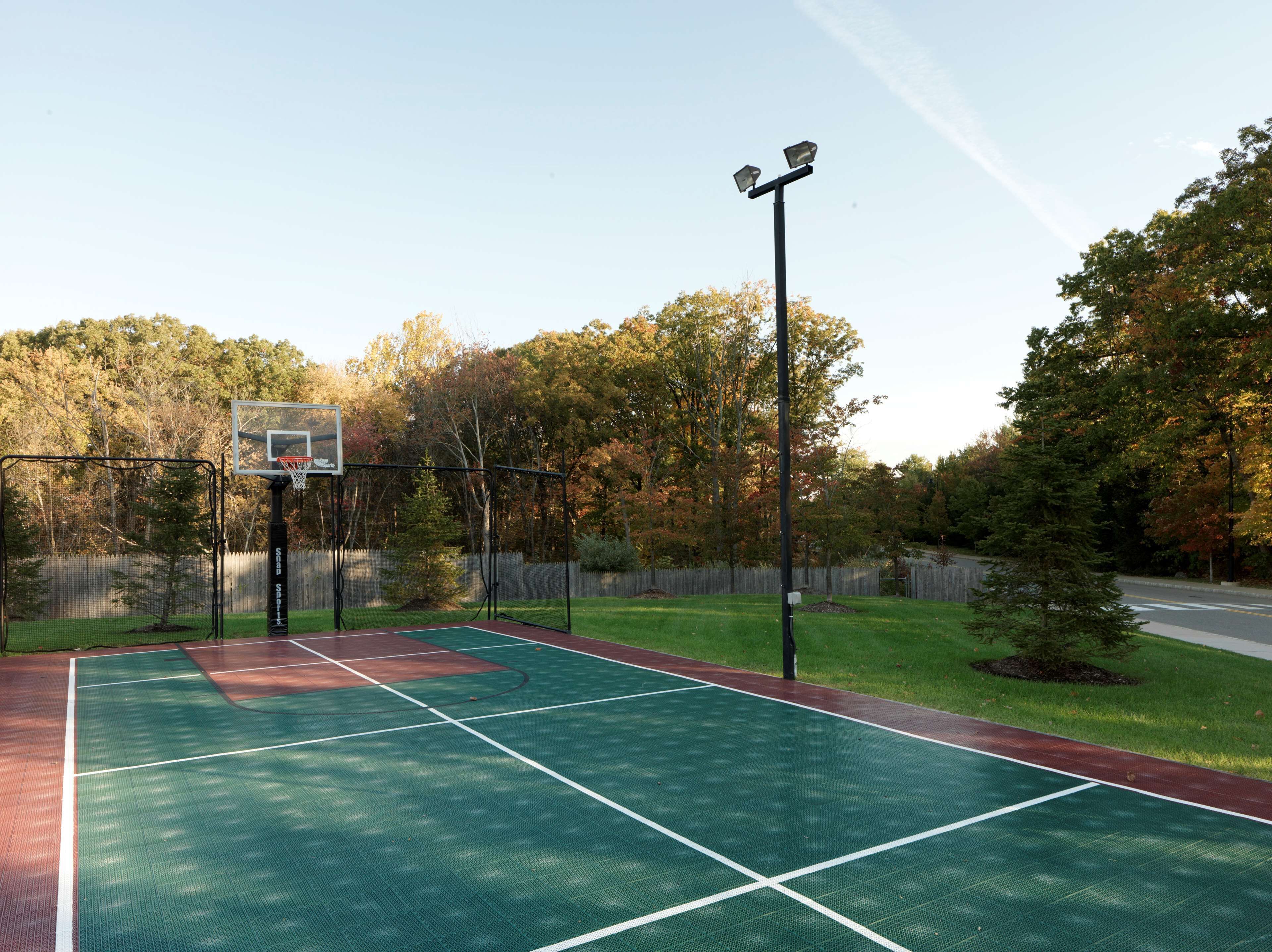 Uma quadra de basquete ao ar livre com cerca, árvores e uma rua ao fundo.