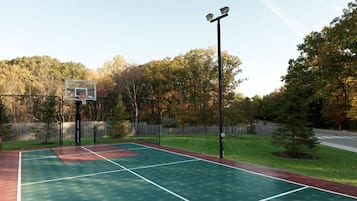 Uma quadra de basquete ao ar livre com cerca, árvores e uma rua ao fundo.
