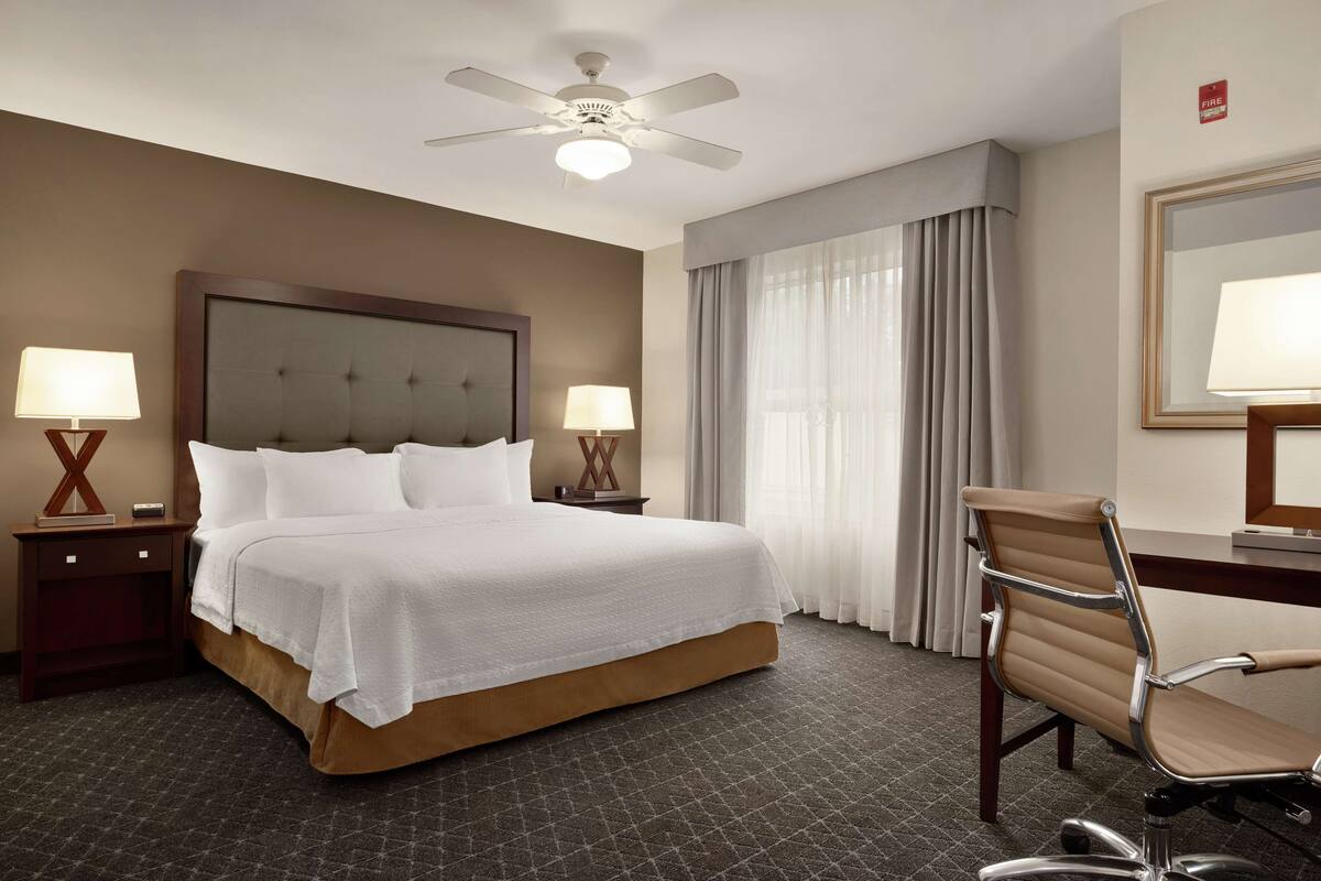 Une chambre d’hôtel avec un grand lit, deux tables de chevet avec des lampes, un bureau avec une chaise, un ventilateur de plafond et une fenêtre avec des rideaux.