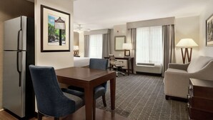 Une chambre d’hôtel équipée d’une table à manger, de deux chaises, d’un canapé, d’un réfrigérateur, d’une lampe et d’un tableau encadré accroché au mur.