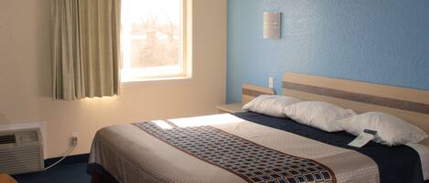 A hotel room with a bed, a window with curtains, a wall-mounted light, and a thermostat.