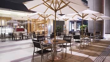 A café with tables and chairs, large umbrellas, and a glass storefront with various items on display.