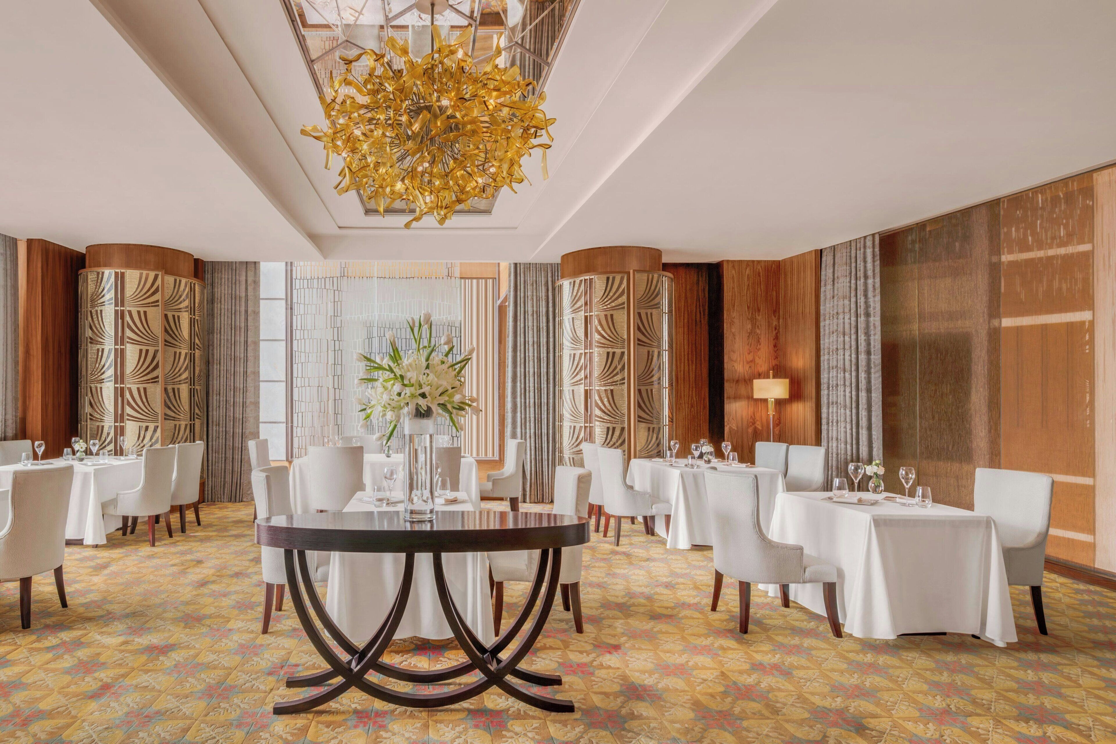 Un comedor de hotel con mesas redondas, sillas blancas y una lámpara de araña central.