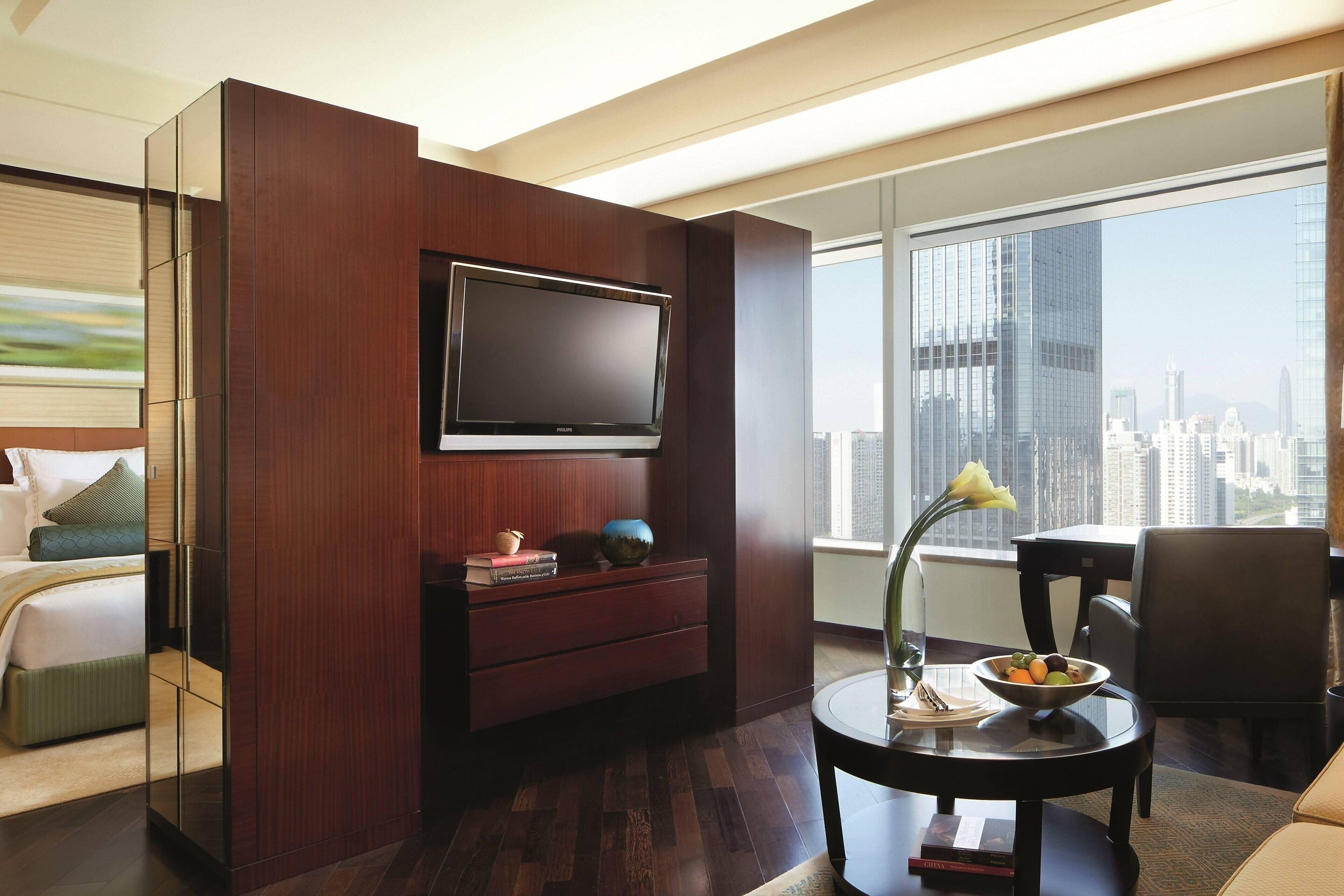 A modern hotel room with a large TV mounted on a wooden panel, a dining area with a table and chairs, and a view of the city skyline through large windows.