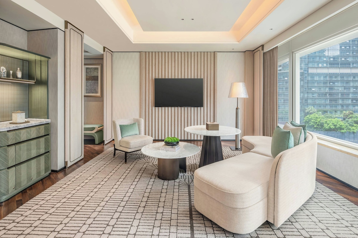 A modern hotel room with a flat-screen TV, a round table, a sofa, and a view of the cityscape.