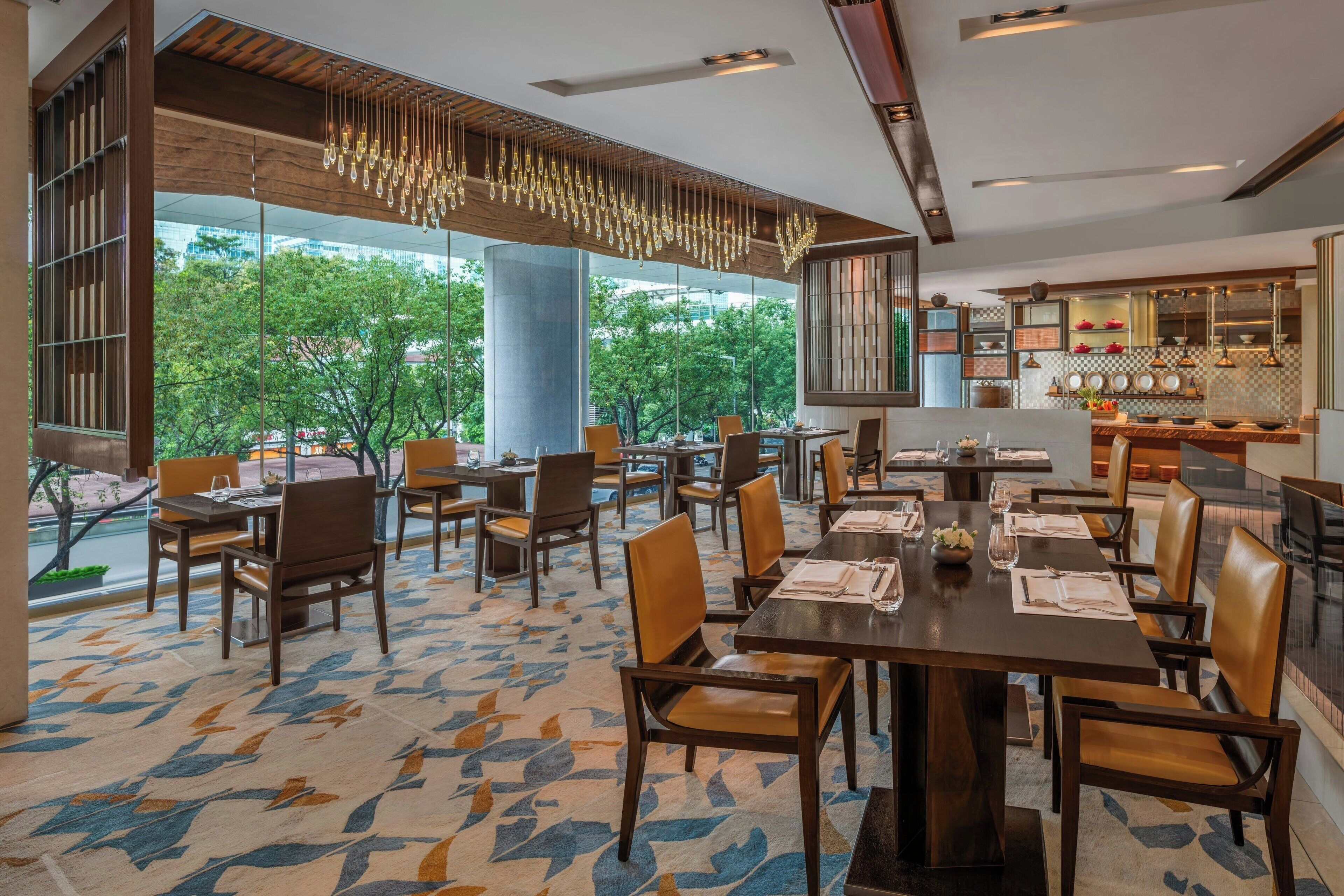 A restaurant with multiple dining tables and chairs, large windows with a view of trees, and a modern chandelier.