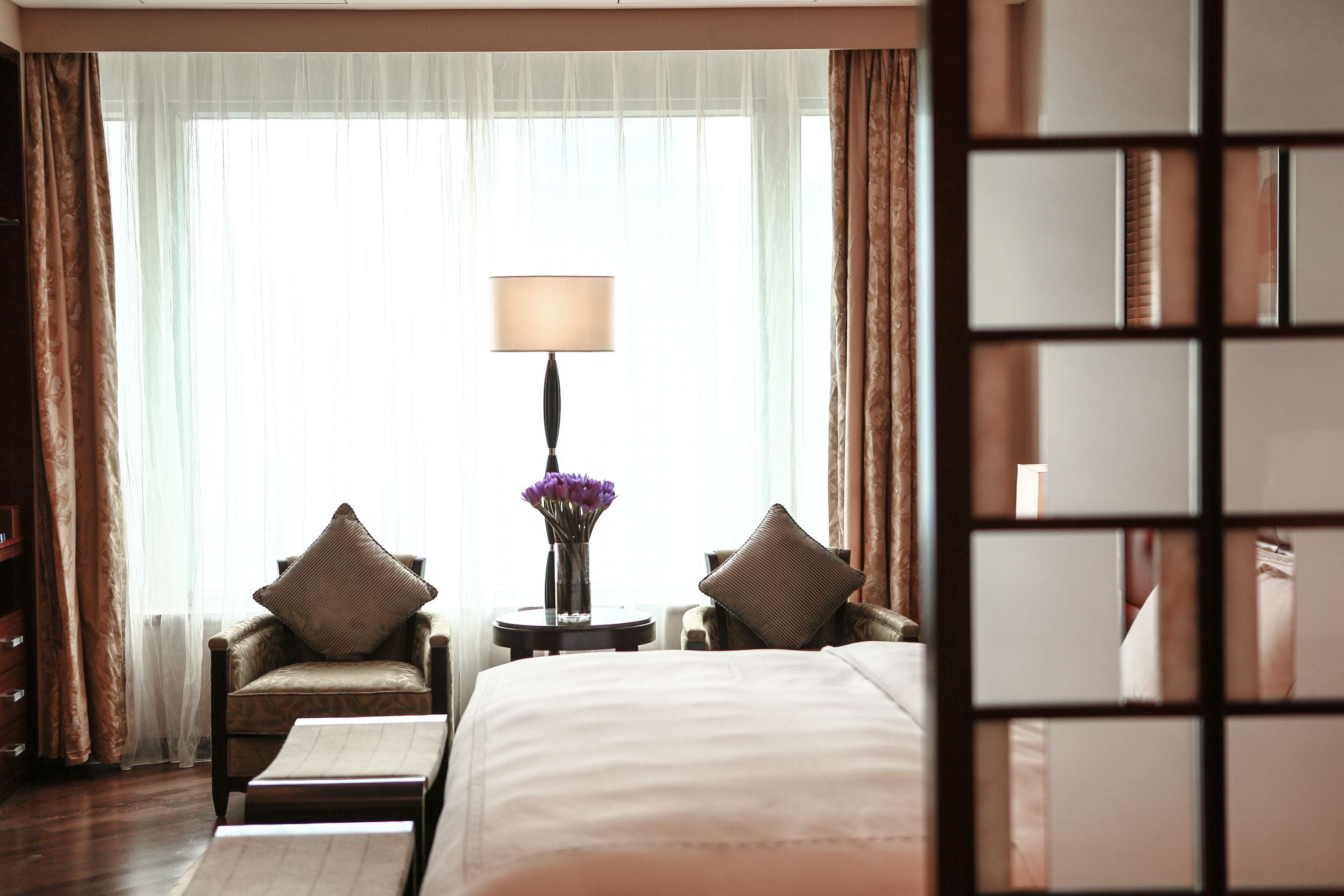 A hotel room with a large bed, two armchairs, a small table with a lamp, and a view through sheer curtains.