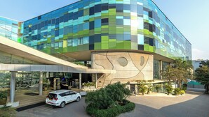 Un edificio moderno con fachada de vidrio curvada, un acceso cubierto y un estacionamiento con un coche blanco.