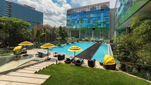Un edificio moderno con fachada de vidrio, piscina con tumbonas y sombrillas, y un césped bien cuidado.