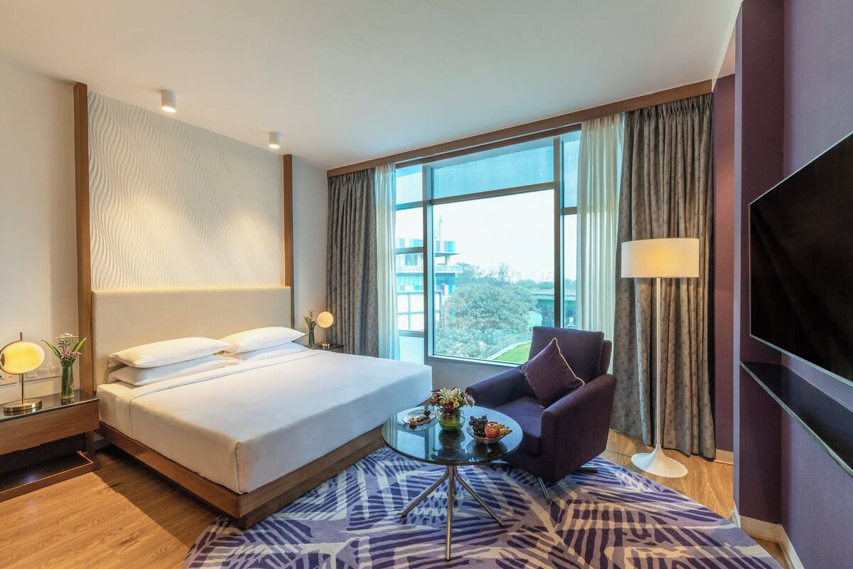 A hotel room with a large bed, a sofa, a small table with a vase and fruit, a TV, and a window with curtains.