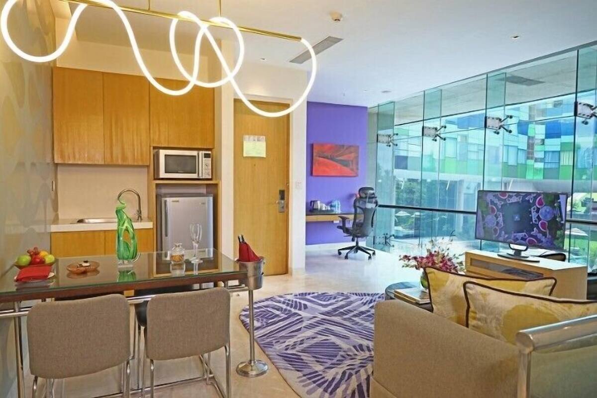A modern living room with a glass wall, a dining area with a glass table, and a kitchen with wooden cabinets and a microwave.