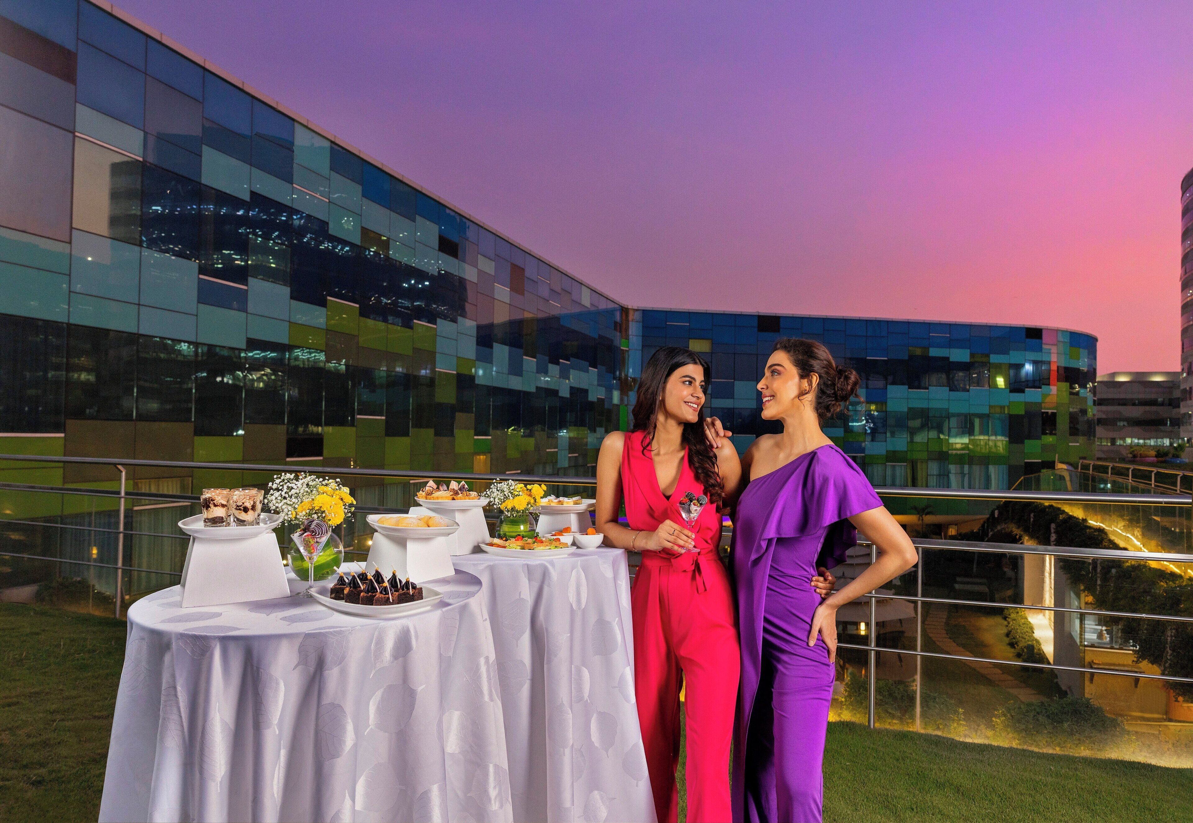 Dos mujeres de pie junto a una mesa con comida y bebidas, con un edificio moderno y un atardecer al fondo.