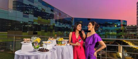 Dos mujeres de pie junto a una mesa con comida y bebidas, con un edificio moderno y un atardecer al fondo.