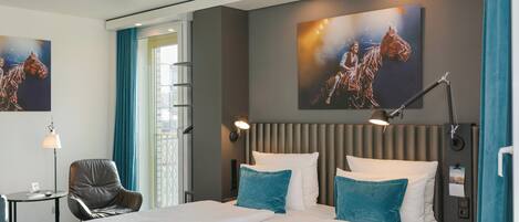 Habitación de hotel con una cama grande, dos almohadas, un escritorio con una silla, una lámpara y obras de arte en las paredes.