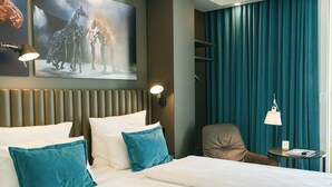 A hotel room with a bed, teal pillows, a headboard, a wall-mounted artwork, a chair, a small table, and a lamp. - Motel One Berlin-Ku'Damm (Berlin)