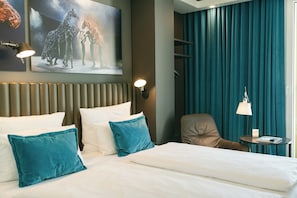 A hotel room with a bed, teal pillows, a headboard, a wall-mounted artwork, a chair, a small table, and a lamp. - Motel One Berlin-Ku'Damm (Berlin)