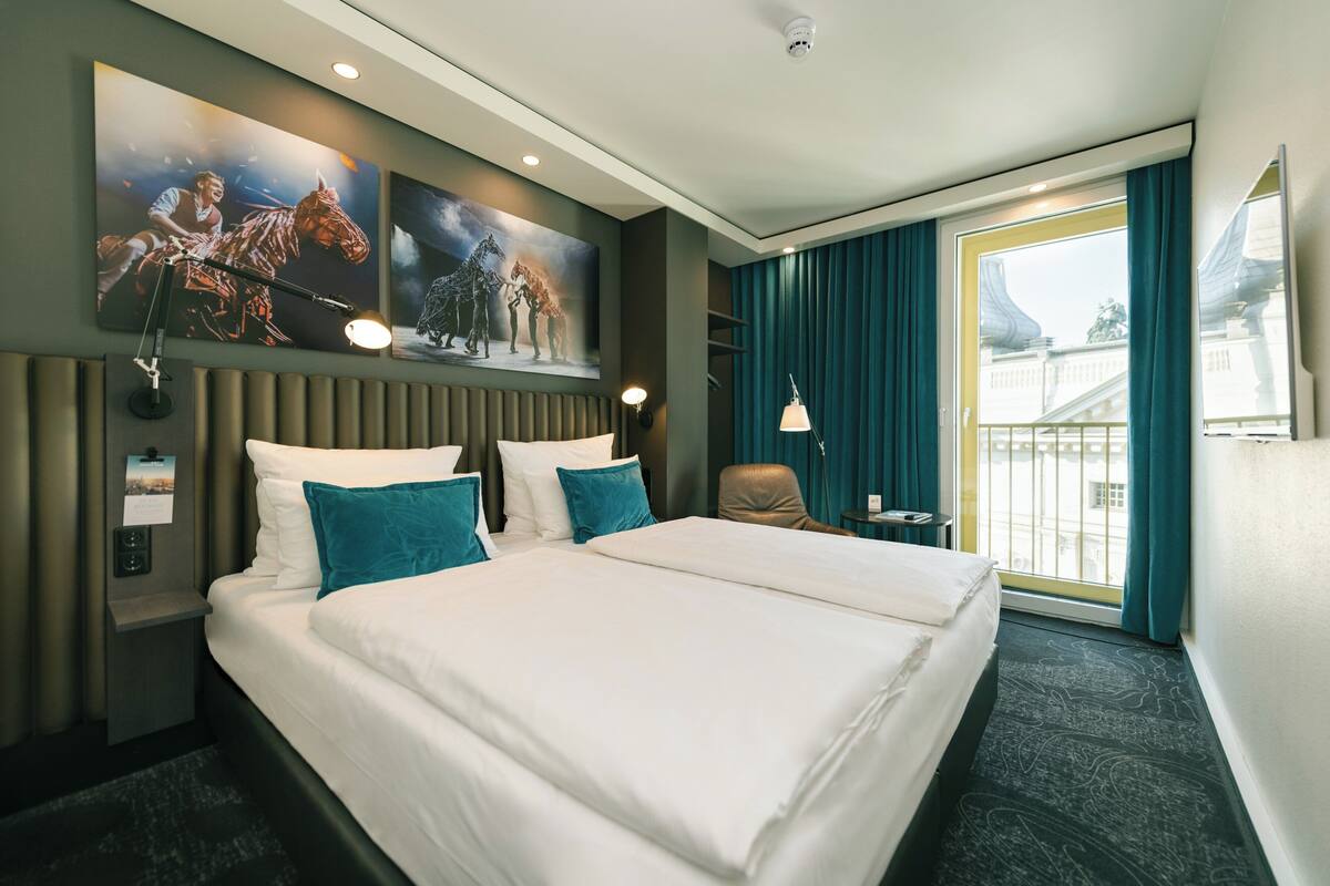 A hotel room with a large bed, a headboard, a desk, a chair, a window with curtains, and artwork on the wall.