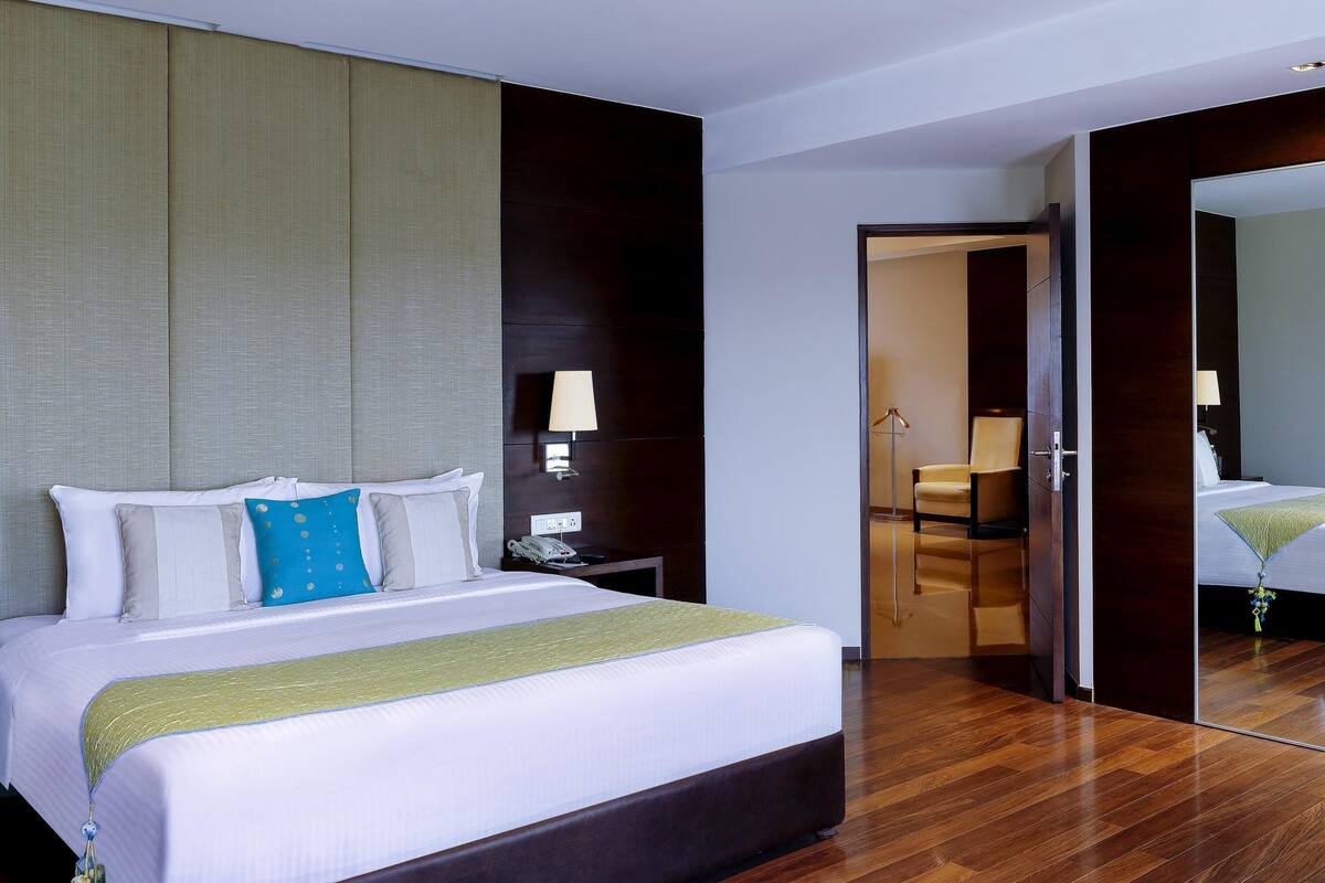 Una habitación de hotel con una cama grande, mesitas de noche, una lámpara, un espejo y una puerta que da a otra habitación.