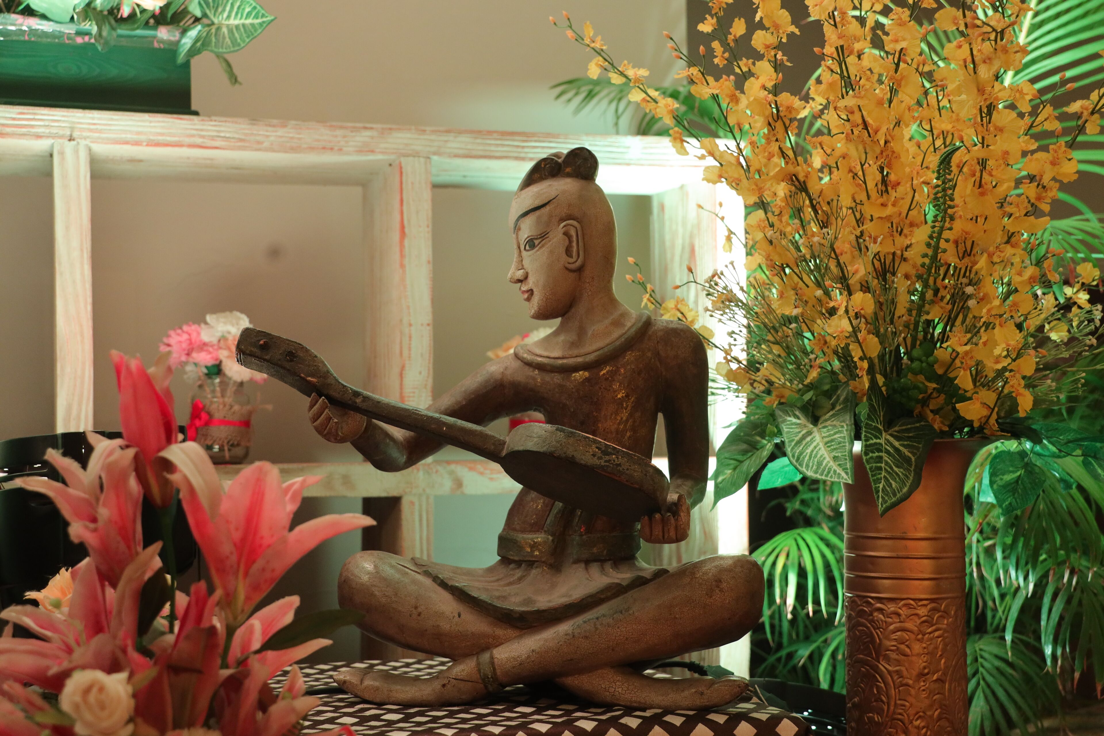 A statue of a seated figure with a long object, surrounded by potted plants and a patterned floor.