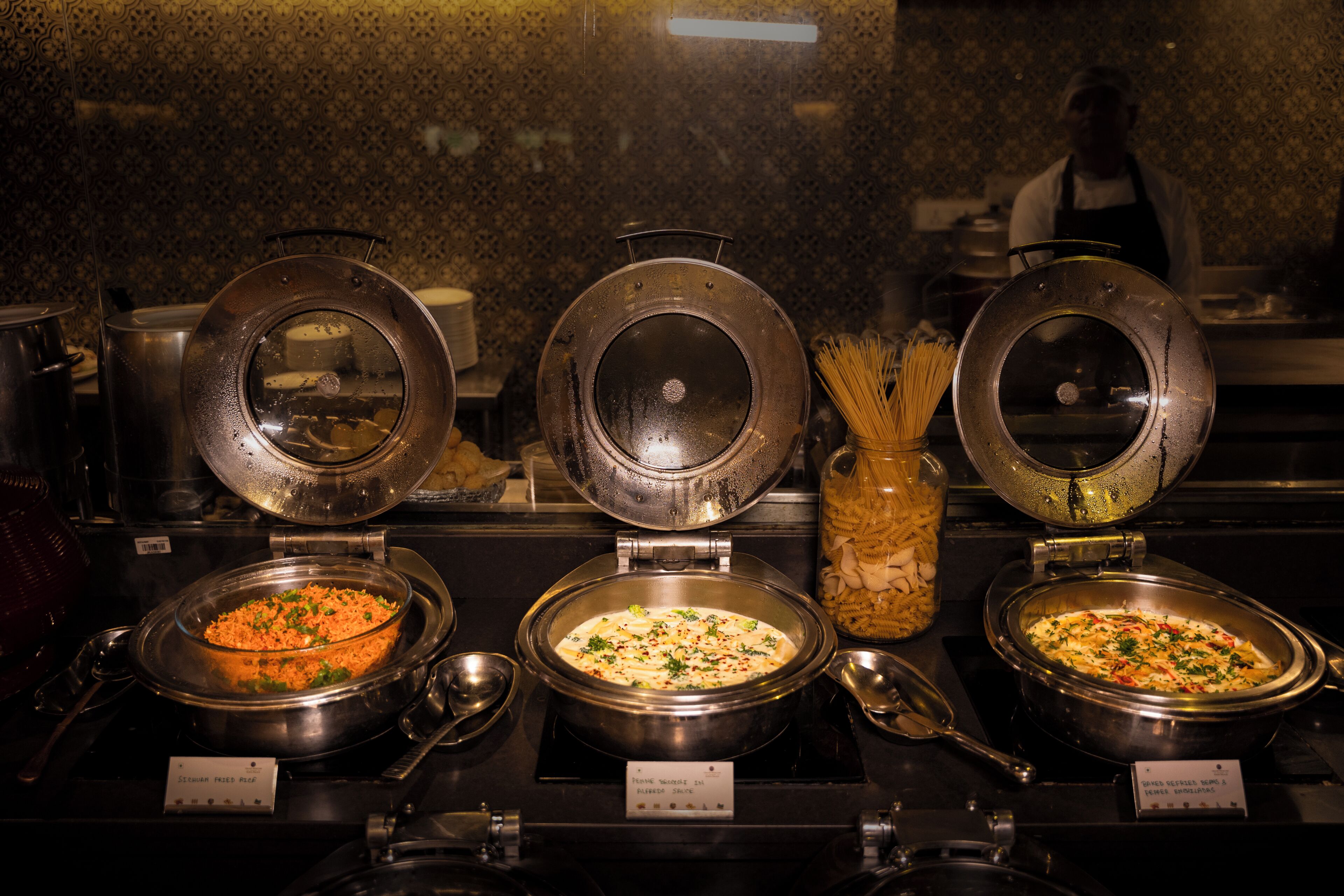 Un bufé con tres recipientes abiertos que contienen diferentes platos, un tarro de pasta y una cuchara.