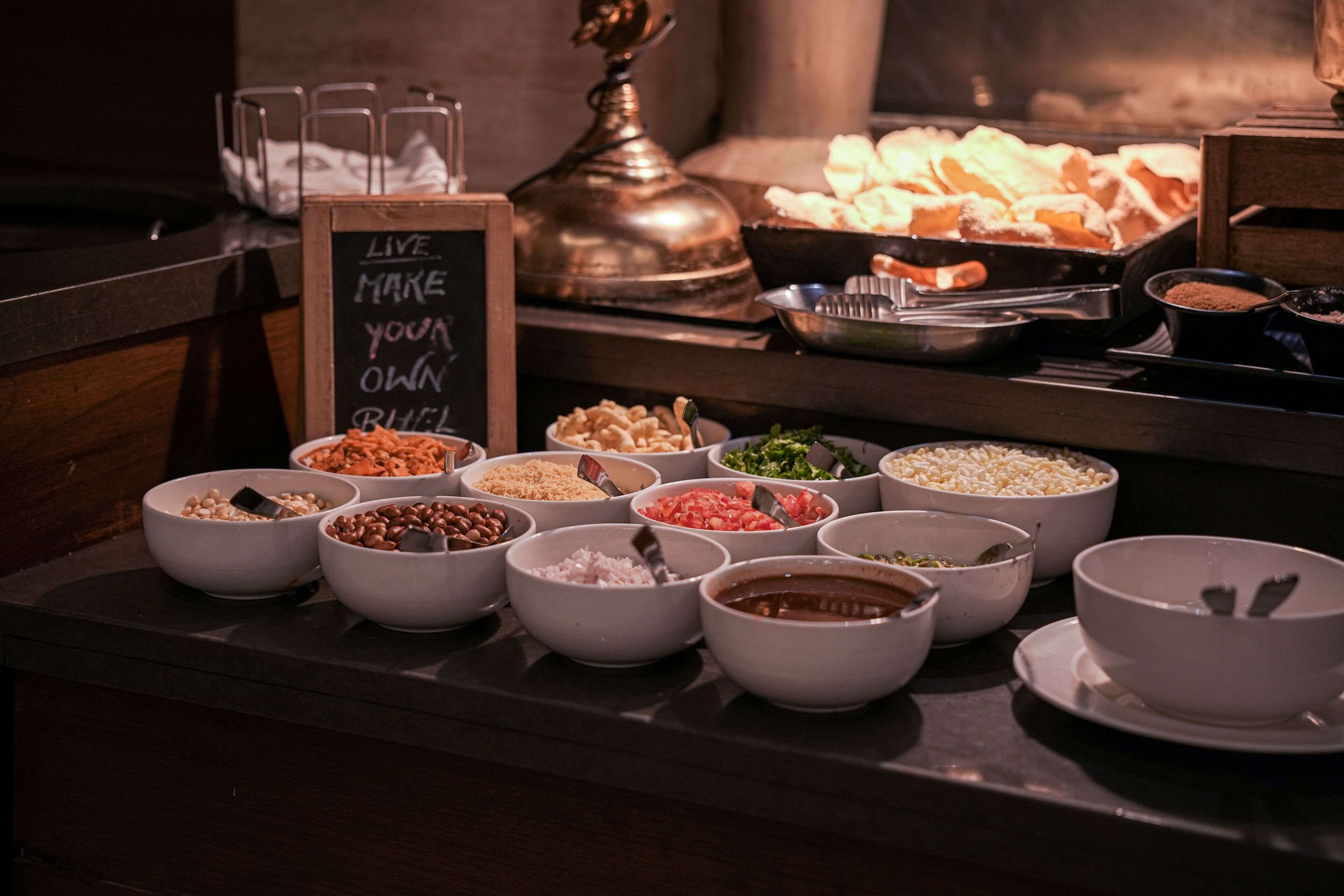 A buffet with various food items in bowls, a sign that says 'LIVE. MAKE YOUR OWN RHEEL,' and a decorative metal object in the background.