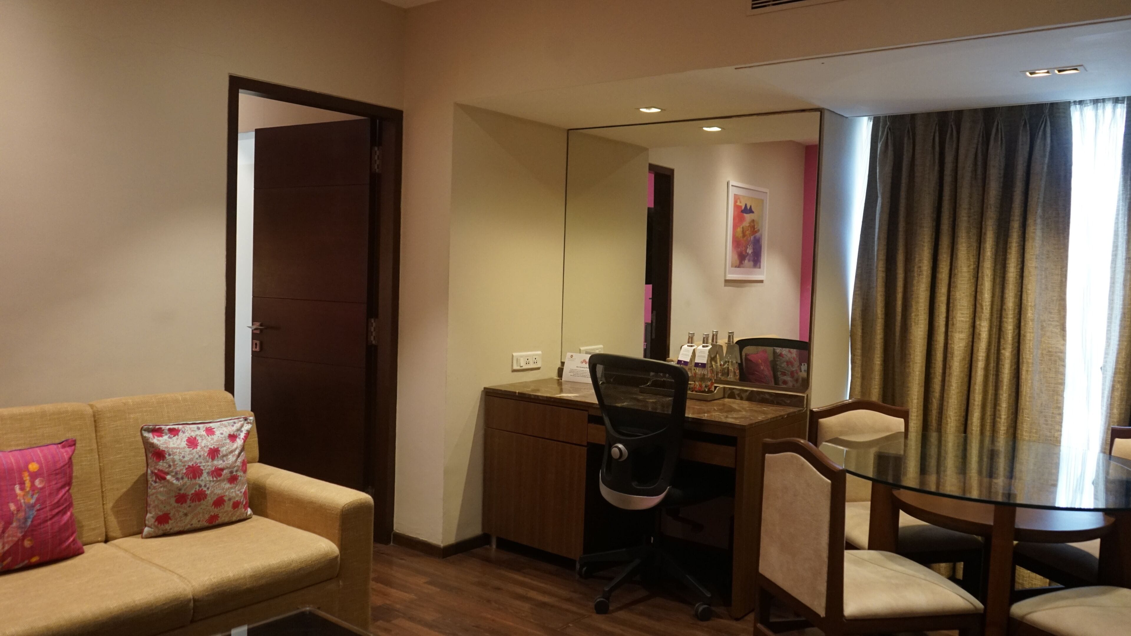 A hotel room with a sofa, a desk with a chair, a dining table with chairs, a mirror, and a window with curtains.