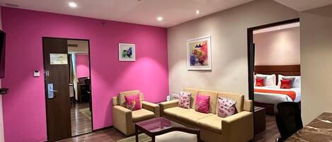A hotel room with a pink wall, a bed, a sofa, a coffee table, and a dining table.