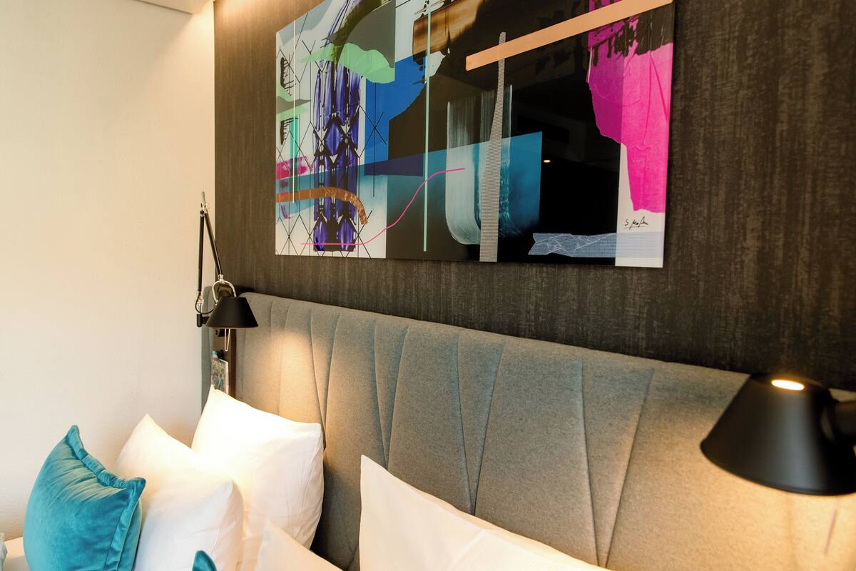 A modern hotel room with a tufted headboard, a bedside lamp, and a colorful abstract artwork on the wall.