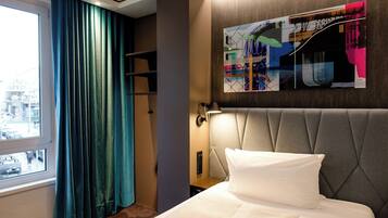 Una habitación de hotel con cama, cortinas en una ventana, una obra de arte en la pared y una mesita de noche con lámpara.