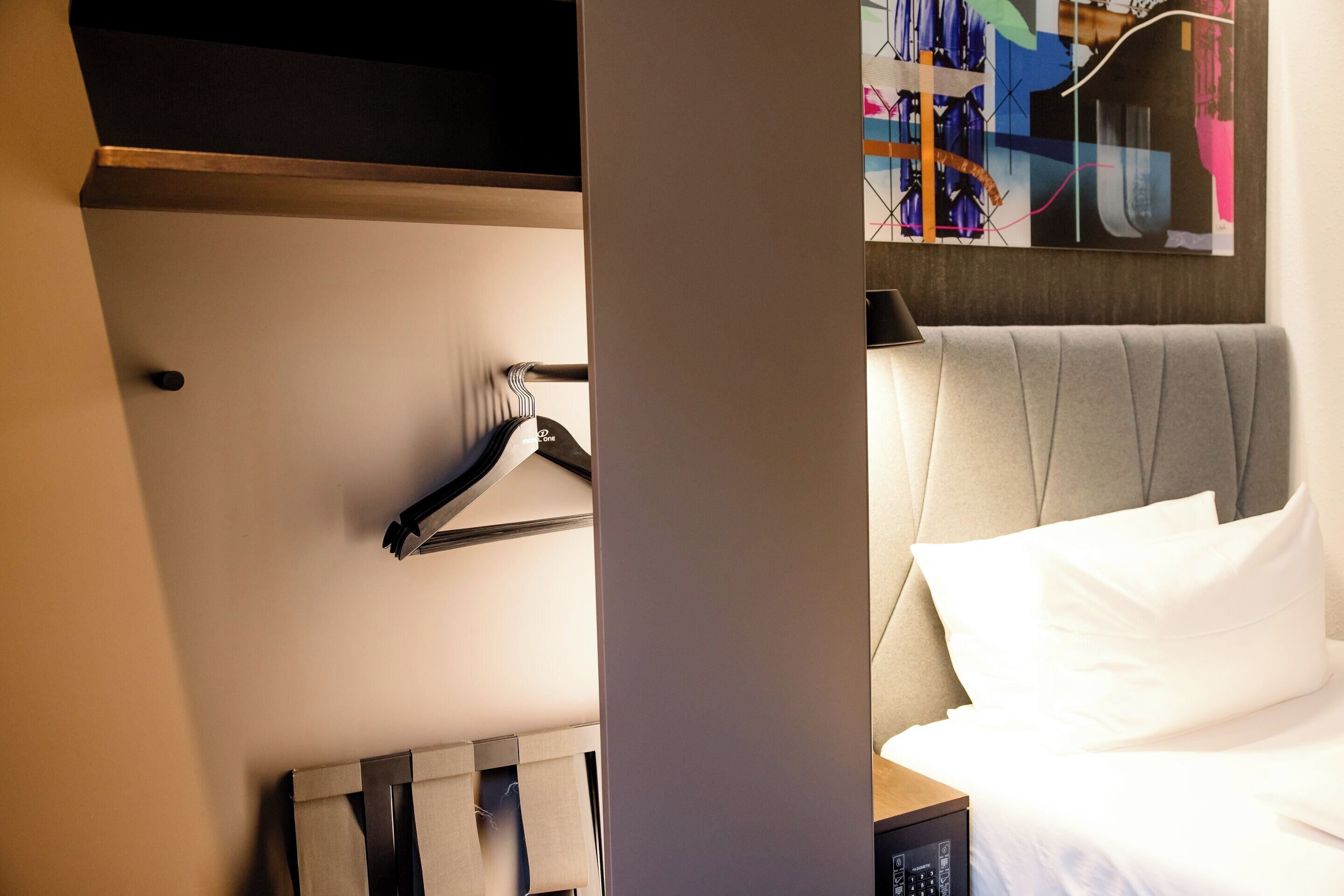 A hotel room with a neatly made bed, a hanging coat rack, and a modern abstract painting on the wall.