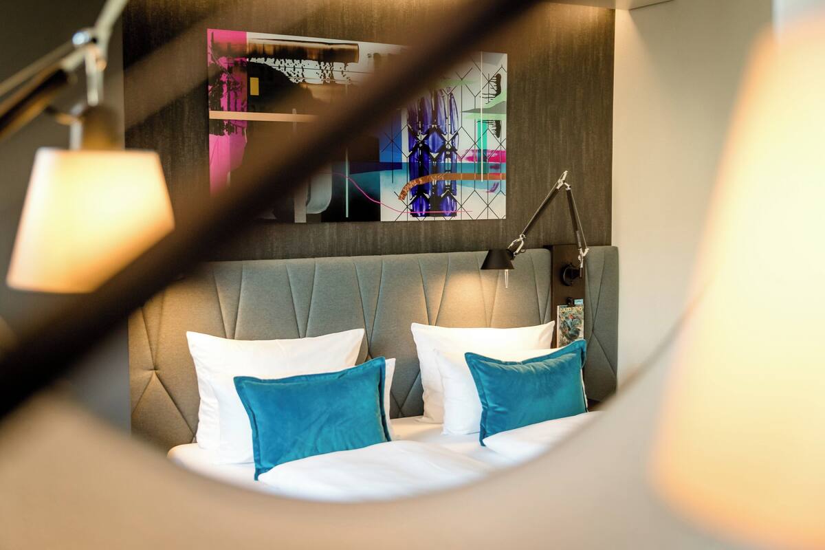 A modern hotel room with a tufted headboard, a large abstract painting on the wall, and a desk lamp.
