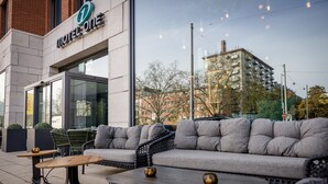 Ett modernt utomhusområde med sittplatser, en soffa och bord, en skylt med texten ”Motel One” och utsikt över byggnader och träd.