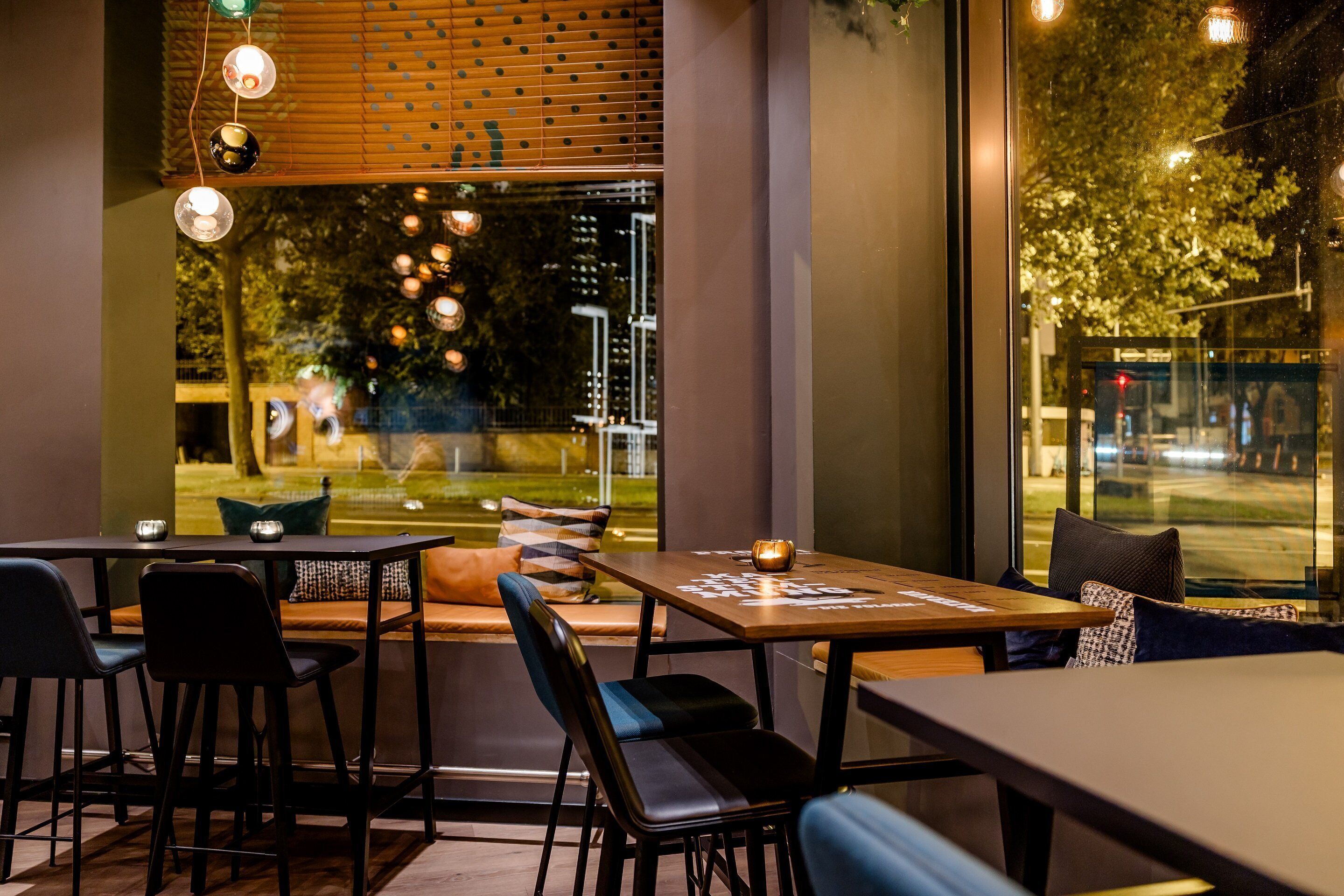 An indoor dining area with tables and chairs, large windows with a view of the outside, and hanging pendant lights.